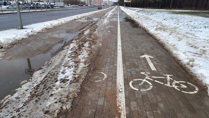 A tile sidewalk and bike path with signage runs along the edge of the city park near trees and cleared of melting snow. Nearby the street roadway passes by and cars are parked Overcast and wet weather