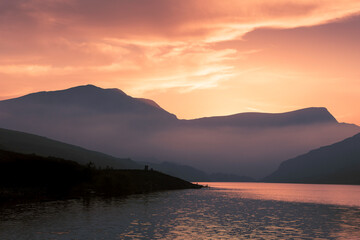 View across Llyn Ogwen lake at sunset, orange sky with mountains silhouette, reflections in the water, campervan and tiny people, Snowdonia, North Wales, UK