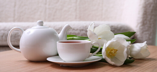 Teapot and cup of hot tea with white tulip flowers on table in living room