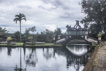 Fototapeta premium Intricate stone bridge over river in Tirtagangga water palace in Bali. Statues, lush vegetation and water fountains.