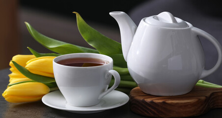 Cup of hot brewed tea, pot and tulips on table against black blurred background