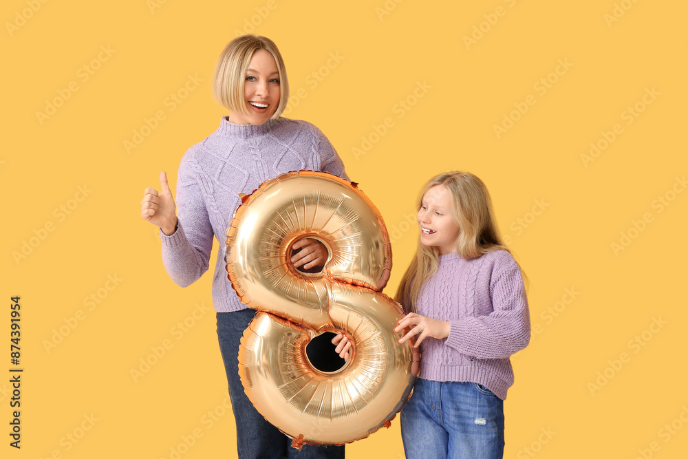 Poster Beautiful young woman showing thumb-up gesture and her cute little daughter with air balloon in shape of figure 8 on yellow background