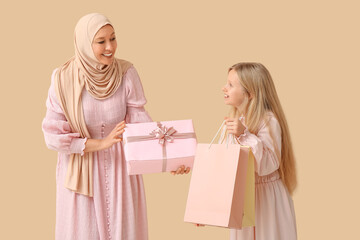 Beautiful young Muslim woman holding gift box and her cute little daughter with paper bags on beige...
