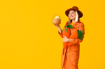 Beautiful young woman in bunny ears with toy carrots and Easter basket on yellow background. Easter celebration