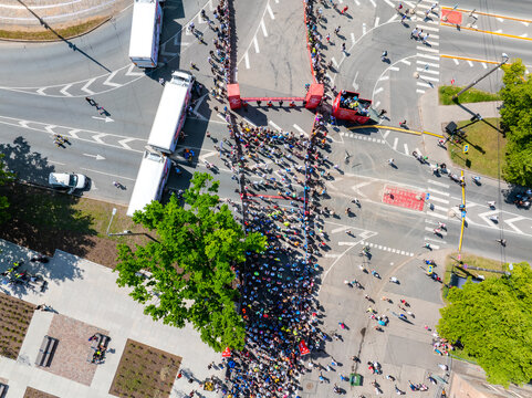 A vibrant aerial shot capturing the energy of a marathon in Riga, Latvia. Runners in bright colors race through city streets, cheered on by enthusiastic spectators.