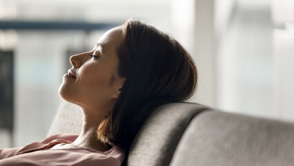 Peaceful beautiful young woman sitting on home sofa with head resting on soft comfortable back,...
