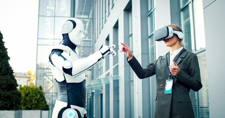 Businesswoman wearing vr-glasses stands in front of robot. Female operates artificial intelligence...