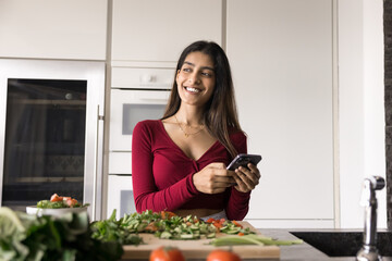 Positive Indian cook girl holding smartphone over heap of organic food on kitchen table, typing on mobile phone, ordering products delivery from grocery store, consulting salad recipe on Internet