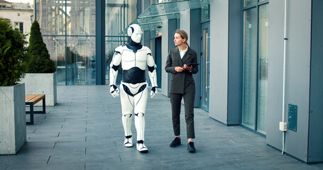Caucasian female wearing business suit goes near building of business center with artificial intelligence machine. Businesswoman speaks with iron robot about work.