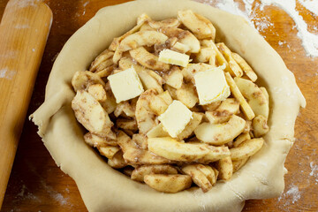 Making a pie.   Rolled dough placed over a pie shell and filled with sliced apple filling with butter.  Top down view.  Rolling pin in background.