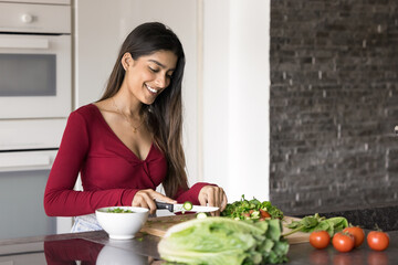 Happy beautiful young adult Indian girl cooking salad in home kitchen, cutting fresh vegetables on board, smiling, enjoying culinary activity, caring for healthy nutrition, organic food diet