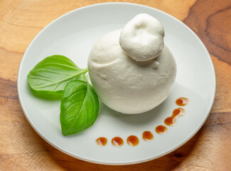 Ball of mozzarella or burrata cheese with fresh basil leaves on white plate on wooden background.