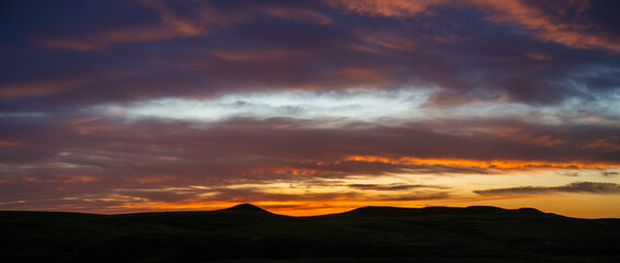 Fototapeta premium Dramatic panoramic view of a multicolored sunset above a silhouette of rolling hills. The sky includes yellow, orange and light purple colored clouds. 
