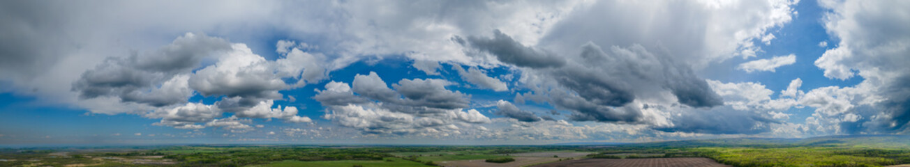 Wide panoramic aerial view of a blue sky with many white summer clouds above a flat agricultural setting.
