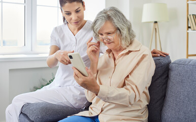Obraz premium Young cheerful female nurse showing elderly woman how to use smartphone. Nursing home service and patient support. Photo of caregiver teaching senior to handle with mobile phone at home.
