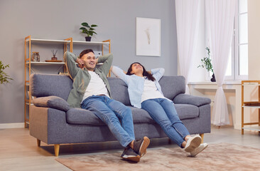 Happy family couple enjoying free time and relaxing on the couch at home. Smiling young man and woman in casual clothes sitting on a comfortable gray sofa in their own apartment. Lifestyle concept