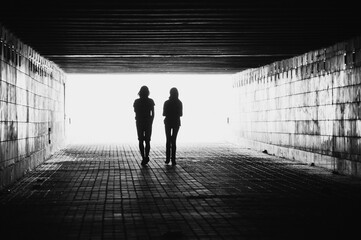 Silhouettes of two people in a tunnel. Black and white image with light coming from behind. Couple walking away from the camera. Lens facing against the light.