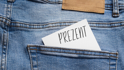 White card with a handwritten inscription "Prezent", inserted into the pocket of blue pants jeasnow (selective focus), translation: A present