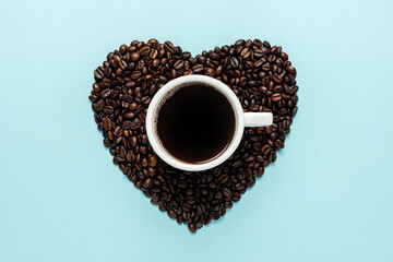 Coffee beans in shape of heart with white cup on blue background. Creative minimal coffee drink concept.