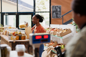 In grocery shop, black woman wearing glasses pours fusilli pasta into bag made of paper. At bio...