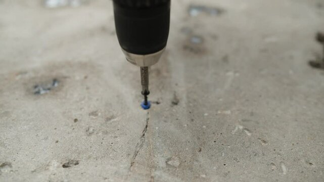 A craftsman screws a self-tapping screw into a concrete floor. A worker repairing a floor. Drive the screw into the concrete floor using a screwdriver.