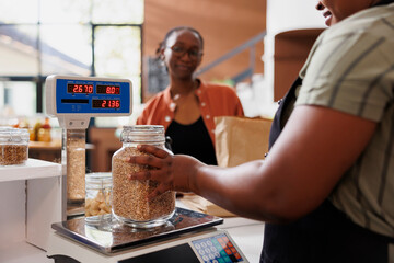 African American woman in an apron assists a customer, weighing locally sourced grains on scale at fresh and organic grocery store. Local merchant measuring jar filled with bio food product.