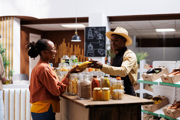 African American male shopkeeper discussing with a female customer about different types of pasta sauce. Image shows young black woman receiving a bottle from vendor in local supermarket.