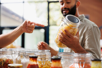 Middle Eastern customer holds and admires a jar filled with organic food products while someone else points at it in a grocery store. Male client analyzing bio locally made item in glass container.