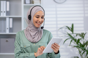 Smiling professional woman wearing headset and hijab using a digital tablet in modern office