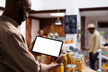 An African American guy consumer holds a phone tablet horizontally with an isolated chromakey mockup template. On wooden shelves, the store sells fresh, eco friendly products.