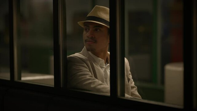 A man wearing a white suit and hat is sitting by a window in a nighttime setting