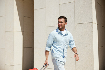 A man in casual light blue shirt and white pants walking with a red suitcase, smiling, outdoors on a sunny day.