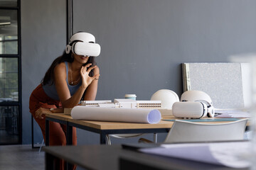 Using VR headset, woman exploring architectural model in modern office workspace, copy space