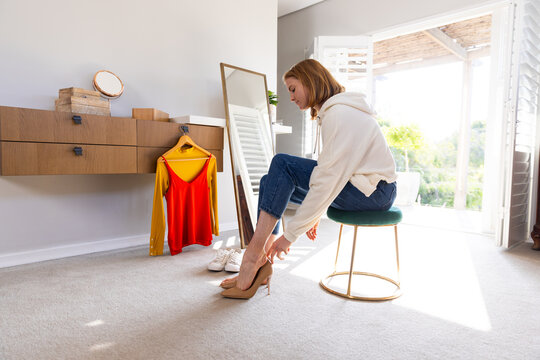 Trying on high heels, woman sitting on stool in bedroom with clothes nearby