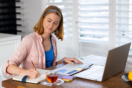 Working from home, woman writing in notepad and using laptop at table - Powered by Adobe