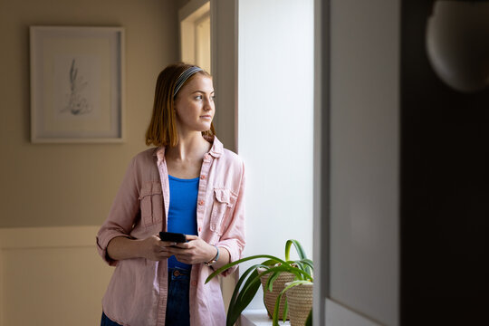 Holding smartphone, woman standing by window and looking outside at home, copy space - Powered by Adobe
