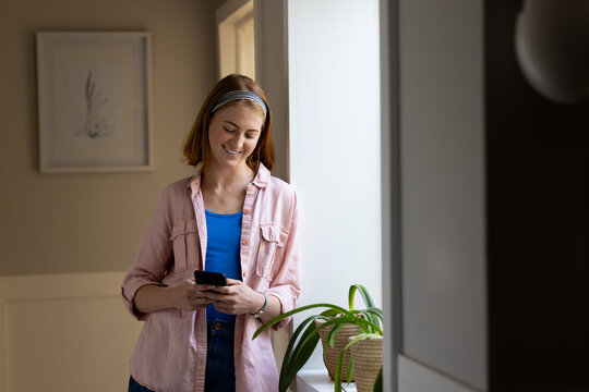 Using smartphone, woman standing by window and smiling in cozy home