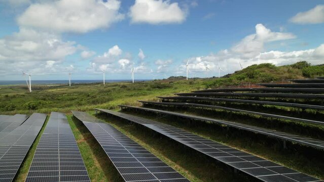 Rows of solar panels with wind turbines being constructed in the distance. Drone video slowly trucking left above solar panels filmed in northern Oahu, Hawaii.