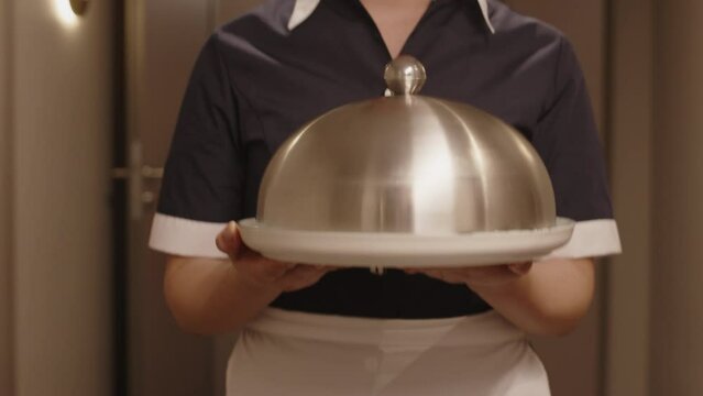 Cropped shot of unrecognizable hotel maid in uniform walking with stainless steel cloche along corridor, delivering food service in room