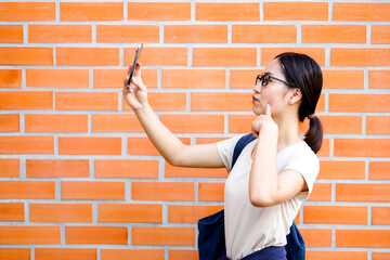 Closeup happiness cheerful Asian young female student in casual cloth holding her smart phone and selfie on brick wall background. Asian school concept.