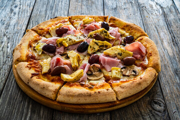 Circle pizza capricciosa slices prepared with cooked ham, mozzarella, artichoke, sliced mushrooms and olives on wooden table

