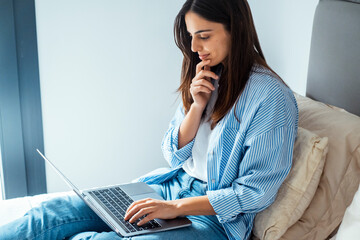 Young adult woman working on laptop sitting on bad with thoughtful expression touching chin and...