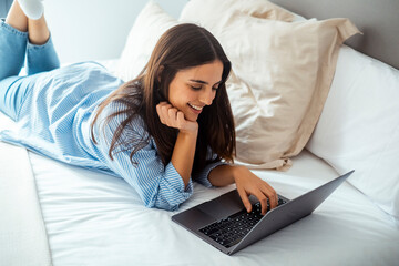Fototapeta premium Lazy modern young woman using social media on laptop laying on bed in morning wake up wearing casual clothes and smiling at the display. Working remote with computer and wireless connection in hotel 