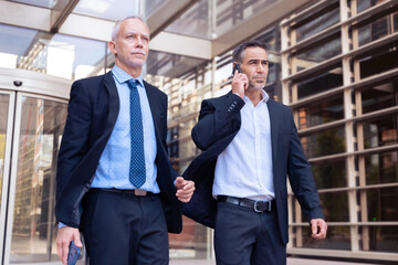 Two Caucasian business colleagues talking on mobile phone, talking coworkers walking around the city outdoors, say goodbye together after work