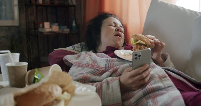 Carefree, careless woman with smartphone and burger licking her lips, yawning. Eaten her fill and want to sleep. Overeating, eat too much food. Unhealthy junk food. Lazy lying on the couch. 