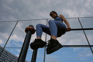woman in blue sportswear performs a pull-up exercise on an outdoor bar against a cloudy sky. This...