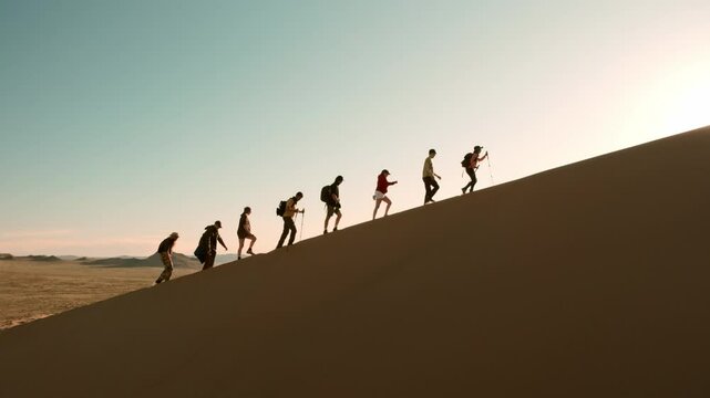 Big group of diverse silhouettes of hikers or tourists are walking uphill on sand dune against sunset sky