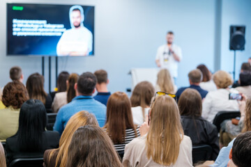 Fototapeta premium Group of people in an auditorium listening to a person giving a presentation at a business conference. Professional setting focused on learning and networking.