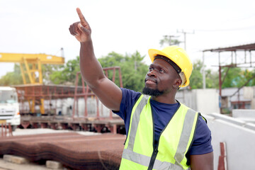 Side view of happy smiling African engineer worker with safety vest and helmet standing and pointing somewhere at construction site. Young architect working at ground level of building site workplace