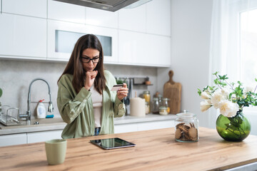 Simple living. Young woman standing in kitchen holding credit card having issue with online payment or shopping on internet with digital tablet. Female banking problems, overspending or finance theft